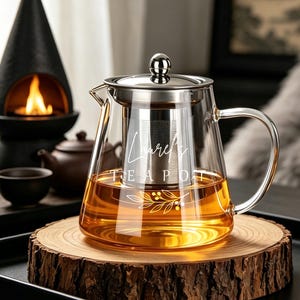 Puede incluir: Una tetera de cristal transparente con tapa de acero inoxidable y un infusor de malla extraíble. El cuerpo de cristal presenta texto blanco en cursiva que dice "Lauren's" sobre "TEA POT" y un gráfico de hojas botánicas.