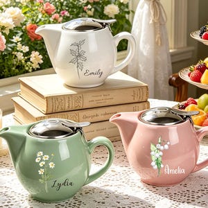 May include: Three ceramic teapots with silver-toned lids. One white teapot features a holly illustration and the name Emily. A sage green teapot displays a daisy graphic and the name Lydia. A pink teapot shows lily of the valley and the name Amelia.