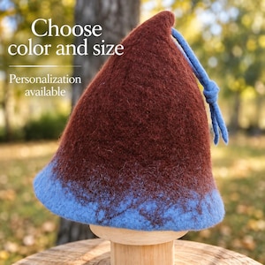 Può includere: Un cappello a punta in feltro con una sezione superiore marrone scuro e una base azzurra. Un cordino azzurro con un nodo è attaccato alla parte superiore. Il testo "Choose color and size" e "Personalization available" è stampato.