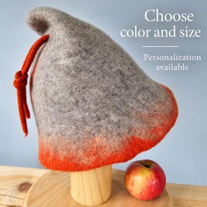 Può includere: Un cappello in feltro grigio con una base sfumata arancione brillante e un cordoncino in feltro arancione abbinato annodato sul lato. Il testo sull'immagine recita: Choose color and size. Personalization available. Una mela rossa e gialla è posizionata accanto.