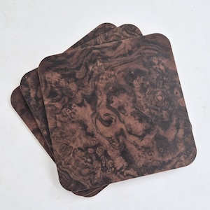 Puede incluir: Un juego de tres posavasos cuadrados con un patrón de veta de madera de color marrón oscuro con texturas orgánicas arremolinadas. Cada posavasos tiene esquinas redondeadas y una superficie plana, dispuestos en una pila ligeramente superpuesta.