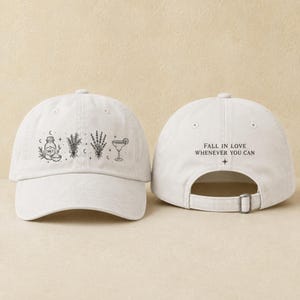May include: A white baseball cap featuring a black line art graphic of a salt shaker, herbs, and a cocktail glass on the front panel. The back panel displays the black text "FALL IN LOVE WHENEVER YOU CAN" with a small four-pointed star symbol below.