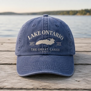 May include: A navy blue baseball cap featuring embroidered text that reads LAKE ONTARIO, EST. 1615, and THE GREAT LAKES, along with a central graphic outline of the lake. The cap has a curved brim, stitched eyelets, and a structured front panel.