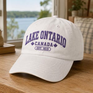 May include: A white baseball cap featuring purple embroidered text that reads LAKE ONTARIO CANADA EST. 1615, accented with two small maple leaf icons. The cap has a curved brim and a structured crown with ventilation eyelets on the top panels.