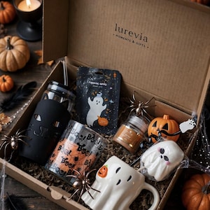 Könnte beinhalten: Eine Papp-Geschenkbox mit der Aufschrift Lurevia Moments & Gifts, die einen Glastrinkbecher mit schwarzem Katzenmotiv, ein Glas mit orangefarbenem und schwarzem Boo-Text, eine geisterförmige Tasse mit Kürbis, eine Snacktüte, eine Kerze und ein Geister-Marshmallow enthält.