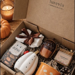 Könnte beinhalten: Eine Pappschachtel mit der Aufschrift lurevia MOMENTS & GIFTS enthält einen weißen Kürbis aus Keramik, einen orangefarbenen Becher mit weißen Kürbisgrafiken und dem Text morning pumpkin, eine braune eichelförmige Kerze, handgemachte Seife und Pralinen.