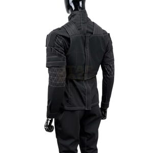 Puede incluir: Una chaqueta t&aacute;ctica negra y pantalones a juego sobre un maniqu&iacute;. La chaqueta presenta un cuello alto, paneles estructurados en los hombros y patrones de costura geom&eacute;trica blanca en las mangas y la espalda. El dise&ntilde;o incluye costuras verticales.