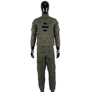 May include: An olive green tactical uniform set consisting of a long-sleeved jacket with a high collar, quilted shoulder panels, and three black rectangular patches on the chest, paired with matching trousers featuring reinforced knee stitching.