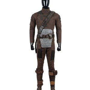 K&ouml;nnte beinhalten: Ein braunes taktisches Kost&uuml;mset bestehend aus einem lang&auml;rmeligen Oberteil und einer passenden Hose. Das Outfit umfasst einen dunkelbraunen Schulterriemen, eine graue Bauchpanzerplatte, einen G&uuml;rtel mit Taschen und braune Beinsch&uuml;tzer.