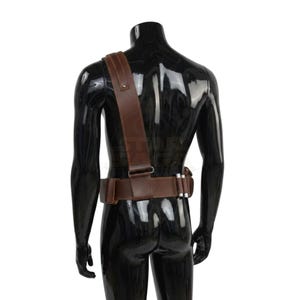 May include: A brown leather utility belt with a matching shoulder strap worn over a black mannequin. The belt features a rectangular pouch, a metal buckle, and three small silver-colored cylindrical canisters attached to the side of the waist.