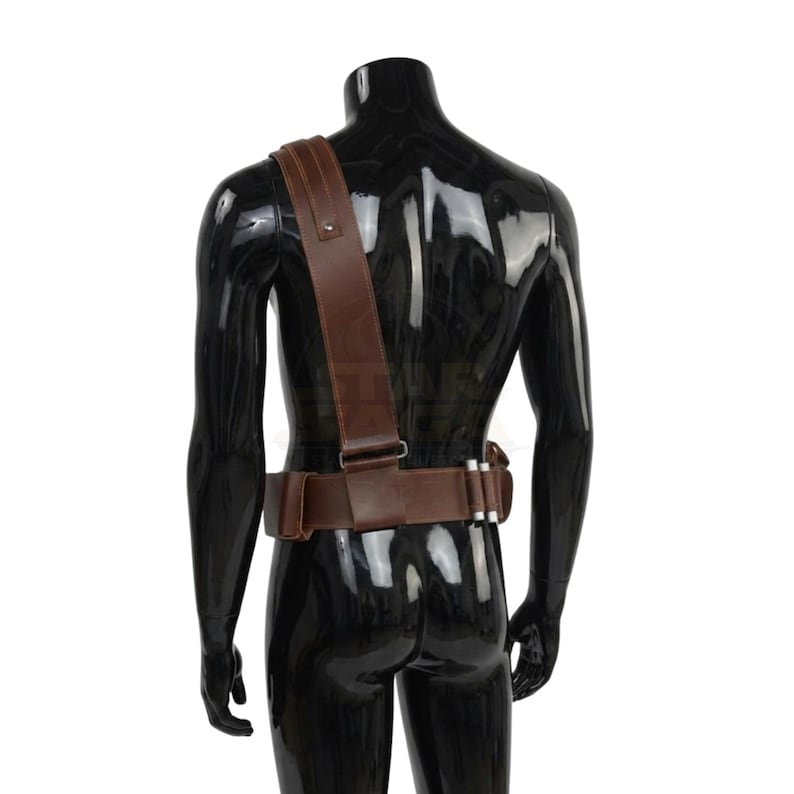 May include: A brown leather utility belt with a matching shoulder strap worn over a black mannequin. The belt features a rectangular pouch, a metal buckle, and three small silver-colored cylindrical canisters attached to the side of the waist.