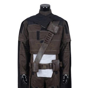 Peut inclure: Un ensemble de costume marron fonc&eacute; et noir comprenant une veste courte avec des coutures horizontales, une cape noire et une ceinture utilitaire marron avec des pochettes et un &eacute;tui. L'ensemble inclut un panneau de taille blanc et une bandouli&egrave;re.