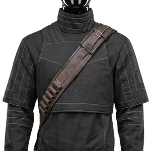 Pode incluir: Uma al&ccedil;a de bandoleira castanho-escura usada na diagonal sobre uma camisa preta de gola alta com costuras brancas. A al&ccedil;a apresenta uma s&eacute;rie de presilhas integradas para segurar pequenos objetos cil&iacute;ndricos, juntamente com fivelas de metal ajust&aacute;veis.