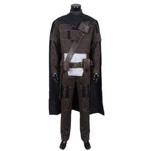 Peut inclure: Un ensemble de costume marron fonc&eacute; comprenant une longue cape noire, un haut marron avec des d&eacute;tails surpiqu&eacute;s et un pantalon marron assorti. La tenue inclut une ceinture utilitaire marron avec des poches et une bandouli&egrave;re diagonale.