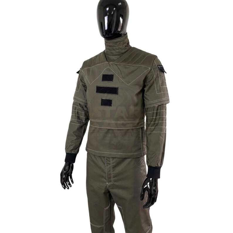 May include: An olive green tactical uniform set featuring a high-neck top with quilted shoulder detailing, white contrast stitching, and black rectangular patches on the chest. The long-sleeved top includes black ribbed cuffs, paired with matching trousers.