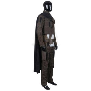 Peut inclure: Un ensemble de costume textur&eacute; marron fonc&eacute; comprenant une longue cape noire, une veste &agrave; col montant avec des d&eacute;tails surpiqu&eacute;s, un pantalon assorti et une ceinture utilitaire avec des pochettes. L'ensemble est pr&eacute;sent&eacute; sur un mannequin noir.