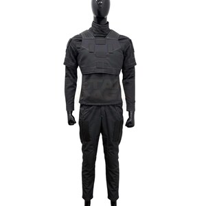 Puede incluir: Un uniforme t&aacute;ctico negro que consiste en una camisa de manga larga con cuello alto, una placa pectoral estructurada con paneles rectangulares y pantalones a juego con grandes bolsillos laterales. El conjunto se muestra sobre un maniqu&iacute; negro.