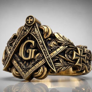 Peut inclure: Une bague en métal doré ornée d'un emblème maçonnique en relief représentant une équerre et un compas avec la lettre G au centre. L'anneau présente des gravures détaillées de feuillage et une petite croix sur le côté avec une patine sombre.