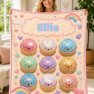 May include: A cream-coloured fleece blanket featuring the name Ellie in blue letters inside a large pink cloud. The design includes nine colourful, glittery, smiling dumpling characters in bamboo steamers, surrounded by rainbows, hearts, and stars.