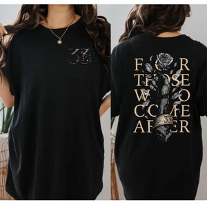 May include: A black t-shirt featuring a small graphic on the front with the number 33. The back displays a large illustration of a hand holding a rose with the text FOR THOSE WHO COME AFTER and the number 33 on a banner wrapped around the wrist.