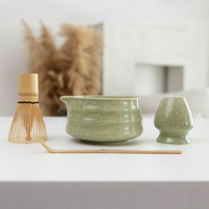 Op de afbeelding: Een matcha-theeset bestaande uit een lichtgroene keramische kom met een tuit, een lichtgroene keramische gardehouder, een bamboe garde met dunne tanden en een lange, slanke bamboe theeschep op een vlakke witte ondergrond.