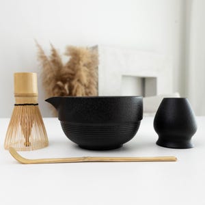 Può includere: Un set da tè matcha composto da una frusta in bambù marrone chiaro, una ciotola in ceramica nera con beccuccio e base scanalata, un supporto per frusta in ceramica nera e un cucchiaio da tè in bambù marrone chiaro lungo e curvo.