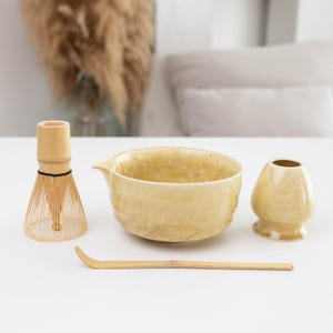 Può includere: Un set da tè matcha composto da una ciotola in ceramica giallo chiaro con beccuccio, un frustino in bambù, un cucchiaino da tè in bambù e un supporto in ceramica giallo chiaro per il frustino. Gli oggetti presentano una finitura smaltata.
