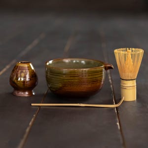 Op de afbeelding: Een matcha theeset bestaande uit een bruin en groen geglazuurde keramische kom met een schenktuit, een bijpassende bruine keramische gardehouder, een lichtgekleurde bamboe garde met dunne tanden en een lange, slanke bamboe theeschep horizontaal.