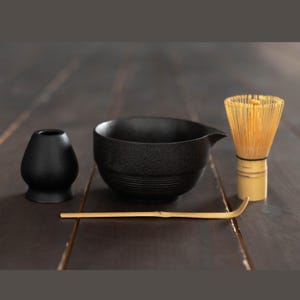 Puede incluir: Un cuenco de matcha de cerámica negra con pico vertedor, un soporte para batidor de cerámica negra, un batidor de bambú de color claro y una cuchara de té de bambú larga, fina y curvada, dispuestos en fila. Objetos de cerámica mate y bambú.