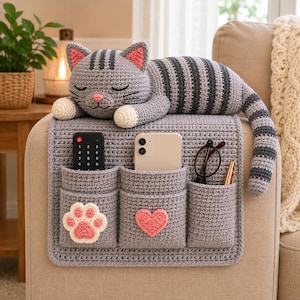 May include: A gray crocheted cat-shaped sofa armrest organizer with dark gray stripes, pink inner ears, and a pink nose. It features three pockets, one decorated with a white and pink paw print and another with a pink heart, holding a remote, phone, and glasses.