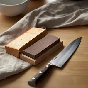 Peut inclure: Un couteau de chef Zwilling avec un manche en bois fonc&eacute; et une lame en acier inoxydable, plac&eacute; &agrave; c&ocirc;t&eacute; d'une pierre &agrave; aiguiser rectangulaire brune sur un socle en bois clair. La pierre est accompagn&eacute;e de sa bo&icirc;te en carton beige TWIN STONE.