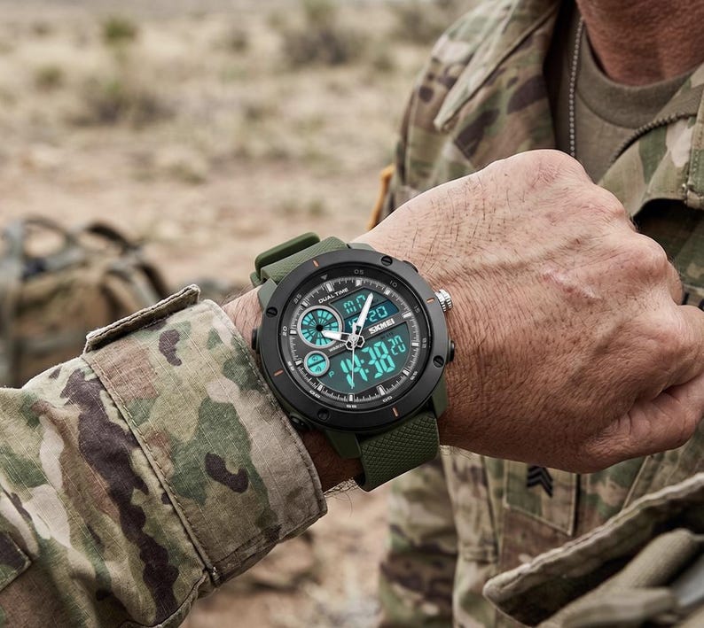 May include: A tactical wristwatch with a matte black circular bezel and a textured olive green silicone strap. The watch face features a dual analog and digital display with the brand name Skmei printed in white, showing a time of 14:38.