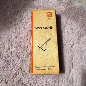 Peut inclure: Une bo&icirc;te rectangulaire de pierre &agrave; aiguiser Zwilling J.A. Henckels Twin Stone avec une finition en grain de bois brun clair. L'avant pr&eacute;sente une bande verticale rouge, une ic&ocirc;ne de couteau noire et le texte TWIN STONE, Wetzstein, Sharpening stone, Pierre &agrave; Aiguiser.