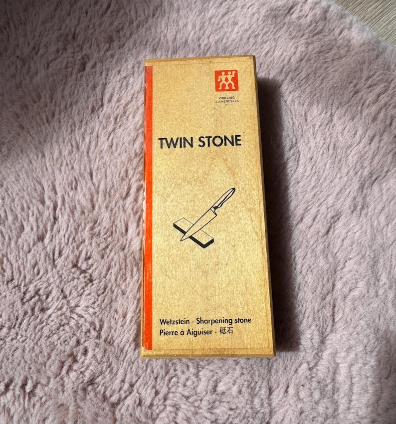 Peut inclure: Une bo&icirc;te rectangulaire de pierre &agrave; aiguiser Zwilling J.A. Henckels Twin Stone avec une finition en grain de bois brun clair. L'avant pr&eacute;sente une bande verticale rouge, une ic&ocirc;ne de couteau noire et le texte TWIN STONE, Wetzstein, Sharpening stone, Pierre &agrave; Aiguiser.