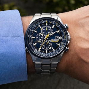 Könnte beinhalten: Eine silberfarbene Edelstahl-Chronographenuhr mit dunkelblauem Zifferblatt, drei Hilfszifferblättern, Datumsanzeige und gelben Akzenten. Das Zifferblatt zeigt den Text World Chronograph WR200 und Blue Angels. Sie hat ein Metallgliederarmband.