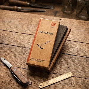 Peut inclure: Une pierre &agrave; aiguiser rectangulaire dans un socle en bois avec un &eacute;tui en carton marqu&eacute; TWIN STONE. L'&eacute;tui pr&eacute;sente un dessin de couteau sur une pierre. Un petit couteau de poche &agrave; manche en bois et une r&egrave;gle en m&eacute;tal sont pos&eacute;s &agrave; c&ocirc;t&eacute;.