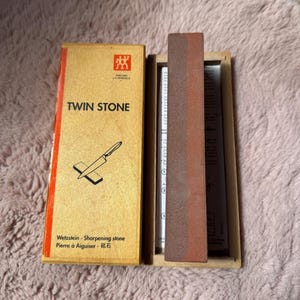 Peut inclure: Une pierre &agrave; aiguiser Zwilling J.A. Henckels Twin Stone dans une bo&icirc;te en bois. Le couvercle beige avec une bande rouge porte l'inscription TWIN STONE et une illustration de couteau. Une pierre &agrave; aiguiser rectangulaire bicolore est &agrave; l'int&eacute;rieur.