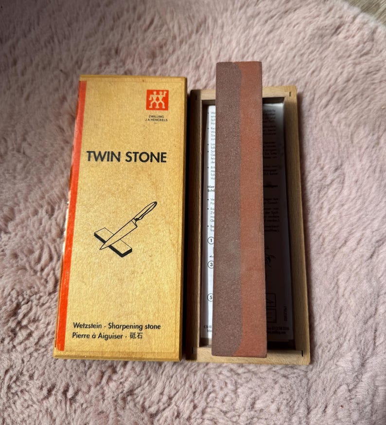 Peut inclure: Une pierre &agrave; aiguiser Zwilling J.A. Henckels Twin Stone dans une bo&icirc;te en bois. Le couvercle beige avec une bande rouge porte l'inscription TWIN STONE et une illustration de couteau. Une pierre &agrave; aiguiser rectangulaire bicolore est &agrave; l'int&eacute;rieur.