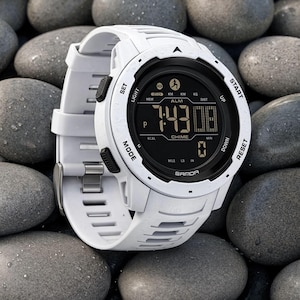 May include: A white digital sports watch with a black circular face and a silicone strap. The display shows the time 7:43, the text SAMOA, and various function labels including SET, LIGHT, MODE, START, RESET, UP, DOWN, ALM, CHIME, KM, KG, and MILE.