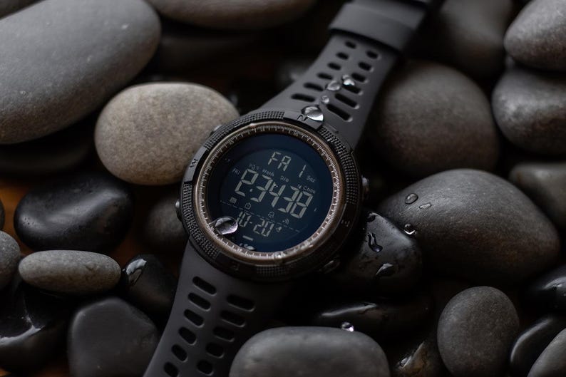 May include: A black digital sports watch with a round face, a textured silicone strap, and a dark display showing the time 2:34:38, the date 10-20, and the day FA 1. The watch features a rugged bezel and is covered in small water droplets.