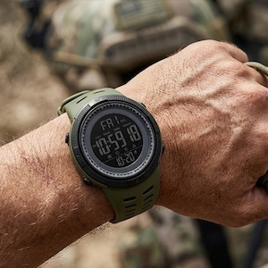 May include: An olive green digital wristwatch with a round black bezel and a textured silicone strap. The digital display shows the day as FRI, the time as 10:59:18, and the date as 10-20, featuring a rugged design with side control buttons.