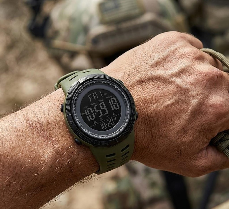 May include: An olive green digital wristwatch with a round black bezel and a textured silicone strap. The digital display shows the day as FRI, the time as 10:59:18, and the date as 10-20, featuring a rugged design with side control buttons.