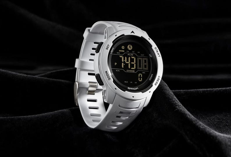 May include: A white digital sports watch with a round face and a black display screen. The watch features a white silicone strap with rectangular cutouts, black side buttons labeled SET, LIGHT, MODE, START, UP, DOWN, and RESET, and the brand name SANDA.