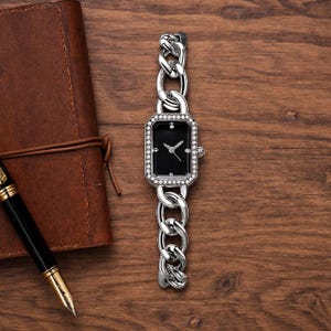 Può includere: Un orologio da polso color argento con quadrante rettangolare nero, lancette argentate e lunetta incastonata di cristalli. Il cinturino è composto da grandi maglie di catena in metallo color argento lucido, creando un aspetto audace.