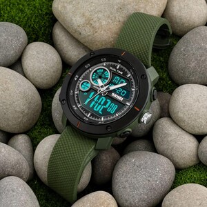 May include: A round analog-digital wristwatch with a matte black bezel and an olive green textured silicone strap. The watch face features a black background with bright teal digital displays, white hour and minute hands, and the brand name SKMEI.