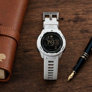 May include: A white digital wristwatch with a black circular face and a silicone strap, positioned next to a brown leather-bound notebook featuring a gold tree emblem and a black fountain pen with gold accents on a dark wooden surface.