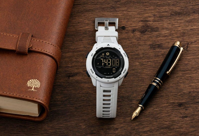 May include: A white digital wristwatch with a black circular face and a silicone strap, positioned next to a brown leather-bound notebook featuring a gold tree emblem and a black fountain pen with gold accents on a dark wooden surface.