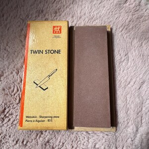 Peut inclure: Un ensemble de pierre &agrave; aiguiser Zwilling J.A. Henckels Twin Stone comprenant une pierre abrasive rectangulaire brune et une bo&icirc;te en carton beige avec une bande rouge, du texte noir et un dessin au trait d'un couteau sur une pierre.