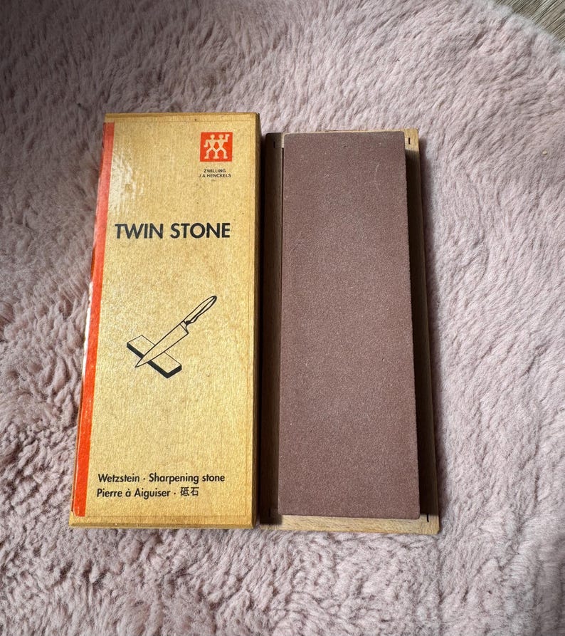 Peut inclure: Un ensemble de pierre &agrave; aiguiser Zwilling J.A. Henckels Twin Stone comprenant une pierre abrasive rectangulaire brune et une bo&icirc;te en carton beige avec une bande rouge, du texte noir et un dessin au trait d'un couteau sur une pierre.