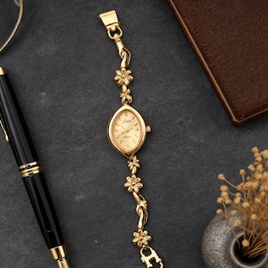 May include: A gold-tone analog watch with a marquise-shaped case and a link bracelet featuring small flower motifs. The watch face is gold with the text "Latt" and "Quartz" printed on it, and it includes a small crown on the right side.